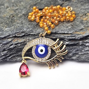 May include: Gold-toned necklace featuring an eye pendant with a blue iris, surrounded by clear crystals. A red teardrop-shaped gem hangs below. The necklace has a beaded chain and is set against a dark, textured background.