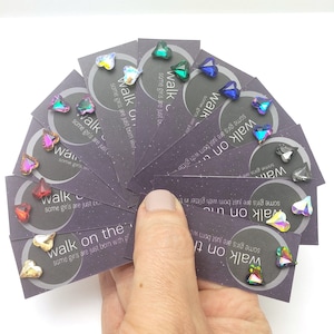 May include: A set of 10 pairs of iridescent heart-shaped earrings. Each pair is displayed on a small card with the text "Walk on" and a quote about girls being born with glitter.