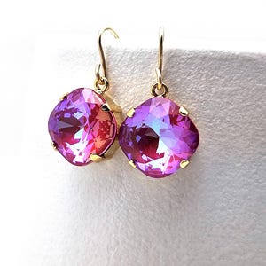 May include: A pair of gold-toned drop earrings featuring large, square-cut, faceted crystals. The crystals transition from pink to purple hues, with a gold setting and a simple hook closure. The earrings are displayed against a white background.