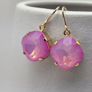 May include: A pair of gold-toned drop earrings featuring large, square-cut, iridescent pink crystals. The crystals are set in gold bezels and hang from delicate gold hooks, creating a glamorous and eye-catching accessory.