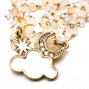 Puede incluir: Un collar de cadena dorada con un dije de nube blanca, un dije de media luna dorada con diamantes de imitación transparentes y un dije de estrella dorada. La cadena está hecha de pequeñas cuentas transparentes y rosadas.