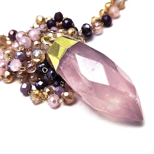 Peut inclure: Un collier avec pendentif en quartz rose, orné d'un cristal facetté et pointu. Le pendentif est coiffé d'un métal doré et suspendu à une chaîne de perles roses, violettes et dorées. Le collier est posé sur fond blanc.
