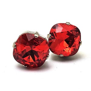 Op de afbeelding: Een paar rode kristallen oorstekers. Elke oorbel heeft een vierkant geslepen, levendig rode kristal, gezet in een zilverkleurige metalen zetting. De kristallen zijn veelzijdig en reflecteren licht, waardoor een sprankelend effect ontstaat.