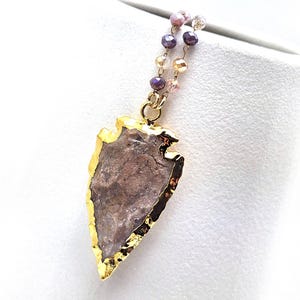 May include: A gold-plated arrowhead pendant necklace with a natural stone center. The stone is a mix of brown and gray hues, and the necklace features a beaded chain with purple, pink, and gold beads. The pendant is approximately 2 inches long.