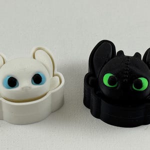 May include: Two 3D-printed figurines of a cartoon dragon. One is white with blue eyes, the other is black with green eyes. Both have large ears and are in a stylized, box-like shape.