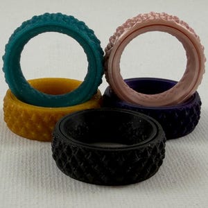 May include: A collection of five circular rings featuring a textured, diamond-patterned exterior. The rings are presented in a variety of solid colors including teal, mustard yellow, light pink, deep purple, and black, arranged in a stacked formation.