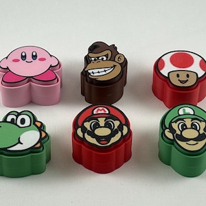 Op de afbeelding: Zes kleurrijke accessoires in de vorm van personages. De personages zijn Kirby, Donkey Kong, een paddenstoel, Yoshi, Mario en Luigi. Elk personage heeft een andere kleur, waaronder roze, bruin, rood, groen en geel. Waarschijnlijk voor een videogame of een gerelateerd product.