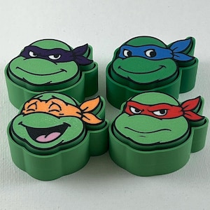 May include: Four green Teenage Mutant Ninja Turtles-themed containers. Each container is shaped like a turtle's head and features a different colored mask: purple, blue, orange, and red. The containers are on a white surface.
