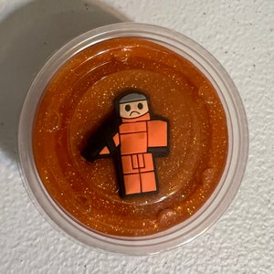 May include: A clear plastic container filled with orange glitter slime with a small orange figurine of a person with a sad face on top.