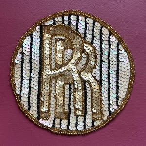 May include: A circular, gold sequined patch with a large, stylized letter "R" in the center. The letter is also made of gold sequins. The background is white and black striped. The patch is surrounded by a gold beaded border, set against a pink background.