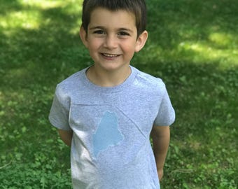 May include: A young boy wearing a grey t-shirt with a blue and white gingham outline of the state of Maine.