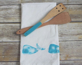 May include: A white kitchen towel with a blue whale design and two wooden kitchen utensils. One utensil is painted blue.
