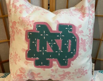 May include: A white pillow with a pink floral pattern and a green and pink felt applique of the letter "ND".