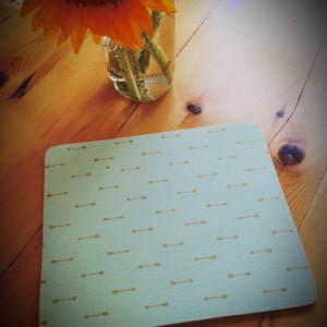 K&ouml;nnte beinhalten: Ein hellblauer quadratischer Platzdeckchen mit einem Muster aus goldenen Pfeilen. Das Platzdeckchen liegt auf einem Holztisch.