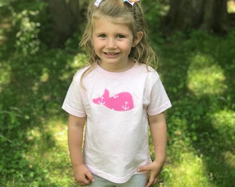 May include: A young girl wearing a white t-shirt with a pink whale applique featuring white anchors. The girl has blonde hair with a colorful bow in her hair. She is smiling and standing in a grassy area.