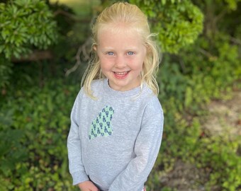 May include: A young girl with blonde hair is wearing a grey sweatshirt with a green and blue lobster design. She is smiling and looking at the camera. She is wearing a skirt with a watermelon print.