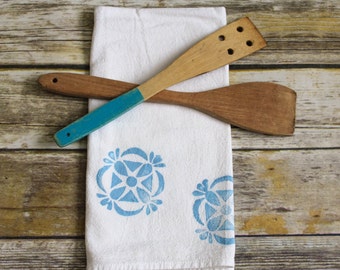 Könnte beinhalten: Zwei hölzerne Küchenspatel, einer in Blau bemalt, liegen gekreuzt auf einem weißen Geschirrtuch mit einem blauen Blumendesign.