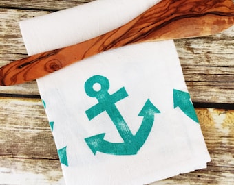 May include: A white kitchen towel with a teal blue anchor design. The towel is folded and a wooden spoon is placed on top of it.