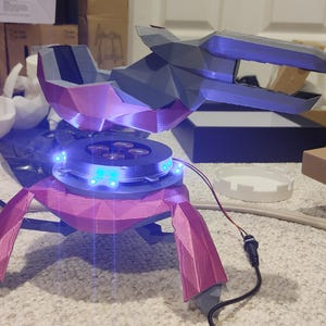 May include: A 3D-printed device in pink and grey, with a futuristic aesthetic. It has a circular central component with blue LED lights and a multi-faceted design. The device has three legs and a top section with a similar geometric design.