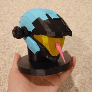 Könnte beinhalten: Ein 3D-gedruckter Helm in Hellblau, Schwarz, Gelb und Pink. Der Helm ist auf einer schwarzen kreisförmigen Basis montiert. Das Visier ist gelb, und eine rosa Zunge erstreckt sich aus dem Mundbereich. Der Helm wird in einer Hand gehalten.