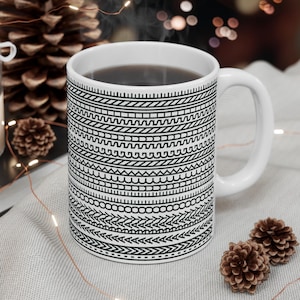 Puede incluir: Una taza de cerámica blanca con un diseño geométrico en blanco y negro, llena de una bebida oscura. La taza está sobre una superficie gris claro, con una vela encendida, piñas y palitos de canela en el fondo.