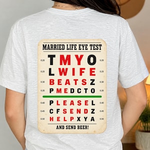 May include: A light gray t-shirt featuring a graphic on the back that mimics an eye chart. The text reads MARRIED LIFE EYE TEST, T M Y O, L WIFE, BEATS Z, P M E D C T O, PLEASE L, C F S E N D Z, H E L P X Y A, AND SEND BEER! with red and black letters.
