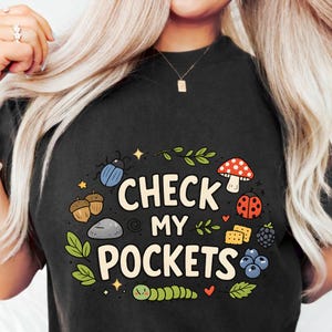 Puede incluir: Camiseta negra con el texto "CHECK MY POCKETS" rodeado de un diseño caprichoso de un escarabajo azul, bellotas, una seta, una mariquita, bayas y una oruga.