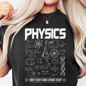 Puede incluir: Camiseta negra con la palabra "PHYSICS" en blanco. La camiseta presenta fórmulas científicas, diagramas y la frase "WHY STUFF DOES OTHER STUFF" en blanco. También se ve un gráfico de un átomo.