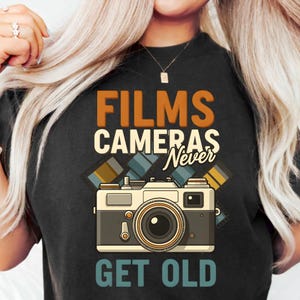 Op de afbeelding: Zwart T-shirt met een vintage camera-afbeelding en de tekst "FILMS CAMERAS Never GET OLD" in oranje en wit. De camera is beige, grijs en zwart met een grote lens. Filmstrips zijn ook afgebeeld.