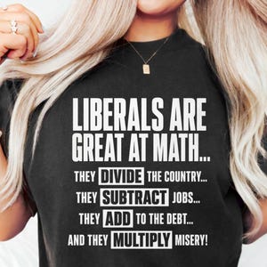 May include: Black t-shirt with white text. The text reads "LIBERALS ARE GREAT AT MATH... THEY DIVIDE THE COUNTRY... THEY SUBTRACT JOBS... THEY ADD TO THE DEBT... AND THEY MULTIPLY MISERY!" The shirt has a crew neck.