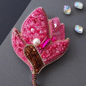 Op de afbeelding: Een roze bloemen broche met bruine en gouden kralen en een parel in het midden. De broche ligt op een grijze achtergrond.