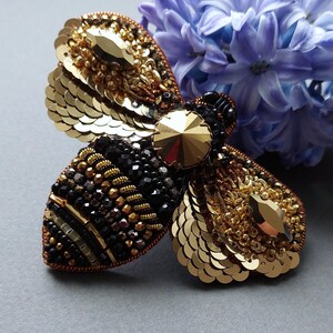 以下が含まれることがあります： クリスタルの中心を持つ、金色と黒のビーズでできた蜂のブローチ。蜂は、スパンコールとビーズで作られており、細部までこだわったデザインです。