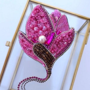 Op de afbeelding: Een roze bloemen broche met een parel en strass, gemaakt met pailletten en kralen. De broche heeft een lange ketting van strass.
