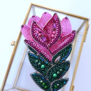 Puede incluir: Un broche de flor de cuentas rosa y verde con lentejuelas y cuentas iridiscentes. El broche se muestra en una caja de vidrio con un marco dorado.
