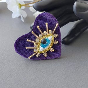 Può includere: Un broche a forma di cuore in velluto viola con un design di occhio malocchio dorato. L'occhio è fatto di perline e paillettes blu e bianche, con un centro nero.
