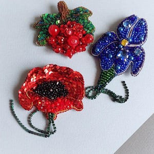 Può includere: Tre spille fatte a mano con perline: un papavero rosso, un grappolo di bacche rosse e un fiore blu. Il papavero ha un centro nero, le bacche hanno foglie verdi e il fiore ha un centro giallo. Il tutto su sfondo bianco.
