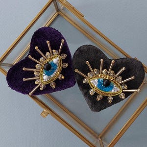 Puede incluir: Dos broches de terciopelo en forma de corazón con un diseño de ojo malvado. Los broches están adornados con cuentas doradas, lentejuelas y pedrería. Un broche es morado y el otro es negro.
