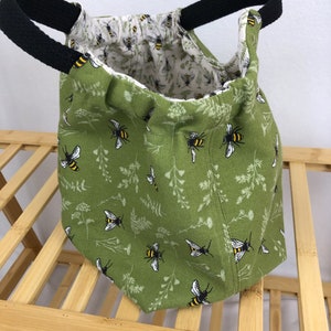 May include: A green fabric tote bag with black handles and a bee and floral print. The bag is empty and sitting on a wooden surface.