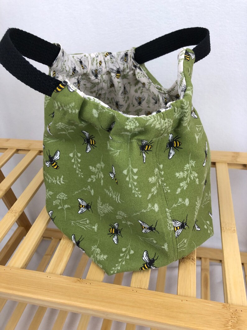 May include: A green fabric tote bag with black handles and a bee and floral print. The bag is empty and sitting on a wooden surface.