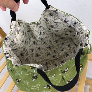 May include: A green fabric tote bag with black handles. The bag is lined with a white fabric with a pattern of bees. The exterior of the bag has a pattern of bees and green foliage.