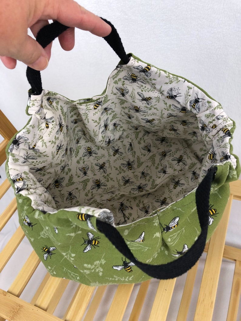 May include: A green fabric tote bag with black handles. The bag is lined with a white fabric with a pattern of bees. The exterior of the bag has a pattern of bees and green foliage.