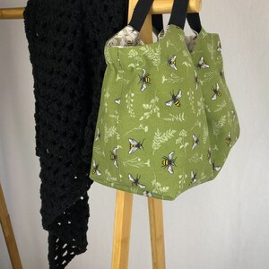May include: A green fabric tote bag with black handles and a bee print. The bag is hanging on a wooden rack.