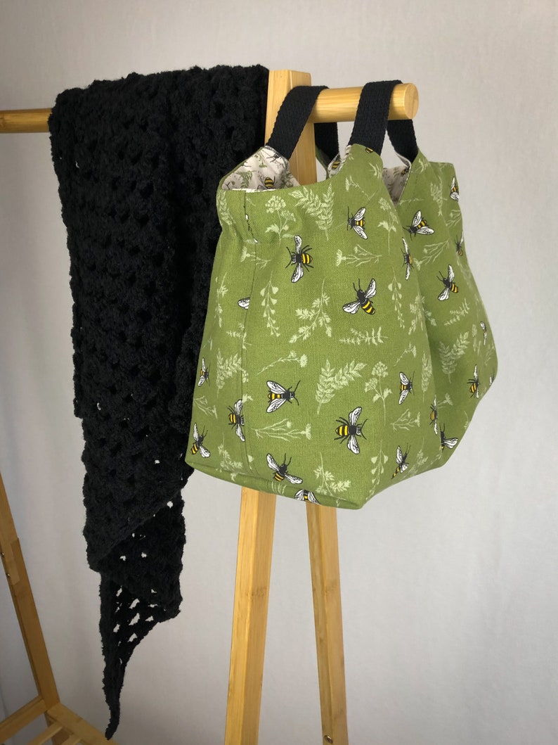 May include: A green fabric tote bag with black handles and a bee print. The bag is hanging on a wooden rack.