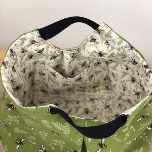 May include: A green canvas tote bag with black handles and a bee print. The bag has a white lining and an interior pocket.