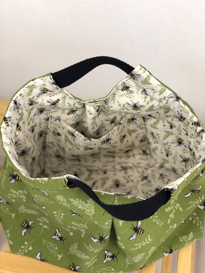 May include: A green canvas tote bag with black handles and a bee print. The bag has a white lining and an interior pocket.