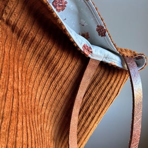 May include: A textured rust-colored corduroy tote bag featuring a light blue interior lining with a repeating dark red floral pattern and two brown leather straps attached to the top edge of the bag.