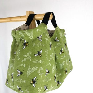 May include: Two green canvas tote bags with black handles and a bee and floral print. The bags are hanging on a wooden rack.