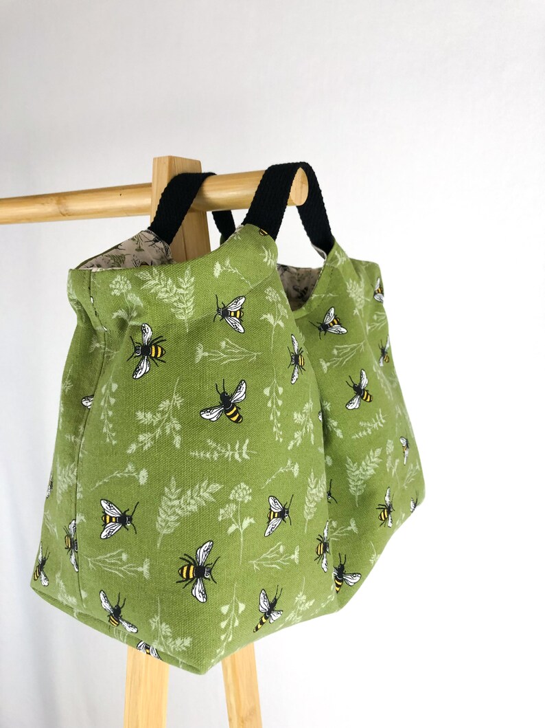 May include: Two green canvas tote bags with black handles and a bee and floral print. The bags are hanging on a wooden rack.