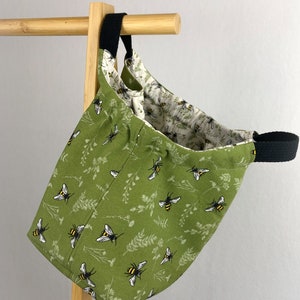 May include: A green canvas tote bag with a black handle and a bee and floral pattern. The bag is hanging on a wooden rack.
