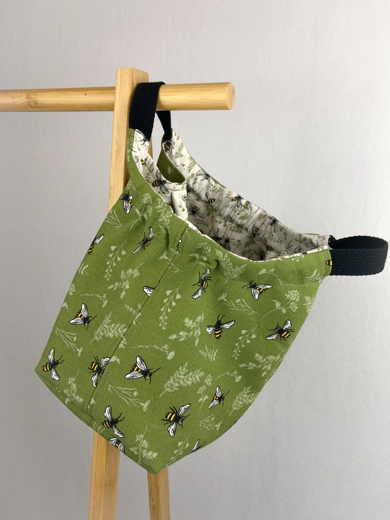 May include: A green canvas tote bag with a black handle and a bee and floral pattern. The bag is hanging on a wooden rack.